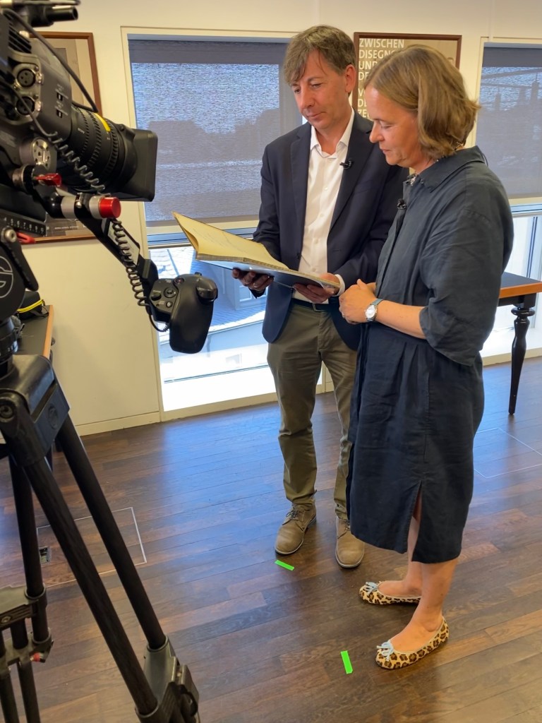 Ein Mann und eine Frau stehen in der Mitte eines Raums und blicken in ein Buch. Vor ihnen steht eine Kamera, die sie filmt. Auf dem Boden sind grüne Markierungen angebracht, die den Personen anzeigen, wo sie stehen sollen.