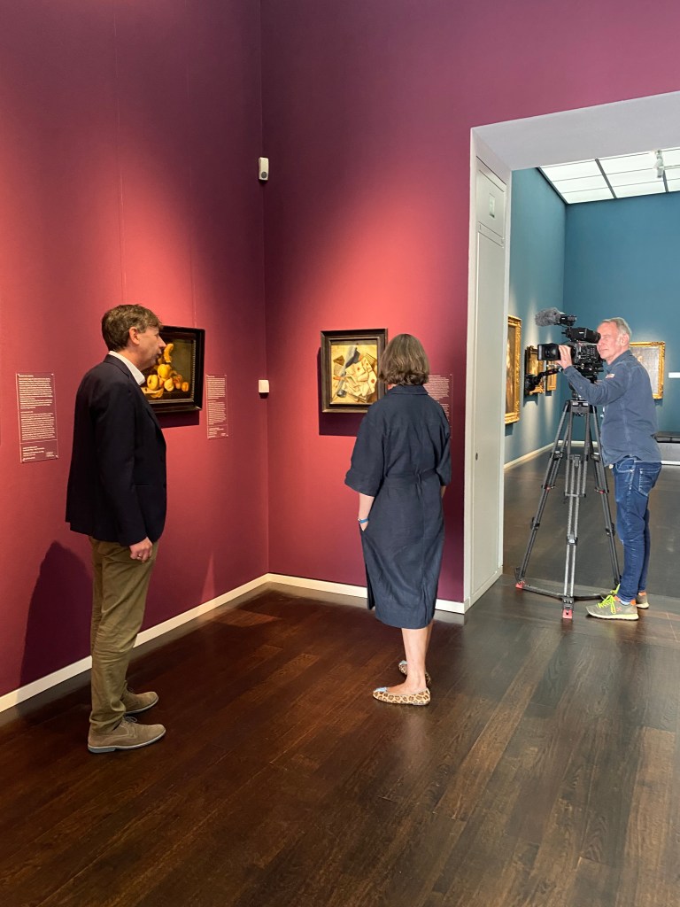 Ein Mann und eine Frau stehen in einem Ausstellungsraum und betrachten ein Gemälde. Rechts neben ihnen im Bild, steht ein Mann mit Filmkamera und nimmt die Gesprächssituation der beiden auf.