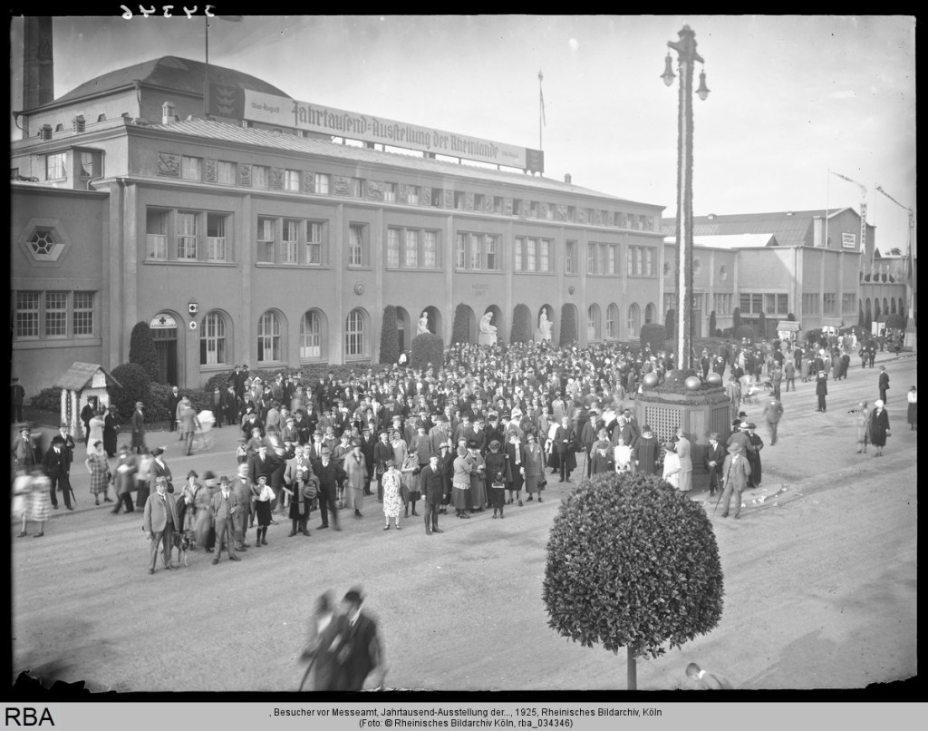 Menschenmenge vor dem Haupteingang der Kölner Messe während der Jahrtausendausstellung im Jahr 1925. Besucherinnen und Besucher aus allen Gesellschaftsschichten versammeln sich neugierig vor dem monumentalen Messegebäude, das mit Fahnen und Schildern geschmückt ist. Die festliche Atmosphäre spiegelt den Stolz auf die regionale Geschichte und den Fortschrittsglauben der Zeit wider.