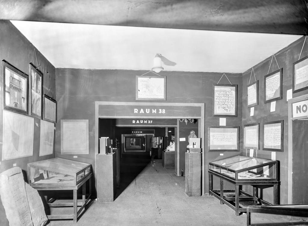 Blick in die Räume 38 bis 36 einer historischen Ausstellung, vermutlich zur jüdischen Kulturgeschichte. Die Wände sind dicht behängt mit hebräischen Inschriften und Zeichnungen. In Vitrinen und auf Sockeln befinden sich weitere Exponate, darunter Inschriftentafeln und Modelle. Die klar strukturierte Raumfolge unterstreicht den musealen Charakter der Präsentation.