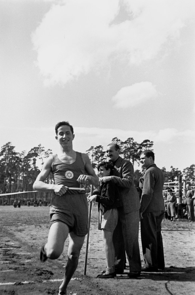 Ein junger Mann läuft durch das Zielband. Auf seinem Trikot ist der Davidstern aufgenäht. Neben der Ziellinie stehen ein Junge und zwei Männer. Im Hintergrund verfolgen weitere Zuschauer*innen den sportlichen Wettkampf.