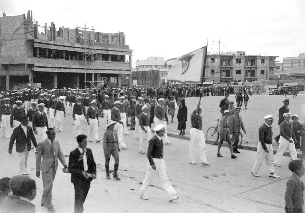 Mehrere Menschen in einheitlicher Kleidung bewegen sich im Bild. Vorneweg läuft ein Mann mit einer weißen Flagge mit Makkabi Logo darauf.